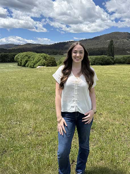 JB Fairfax Award recipient Tayla Gofton, a young woman with long brunette hair wearing casual top and jeans, smiling in a grassy, rural landscape.