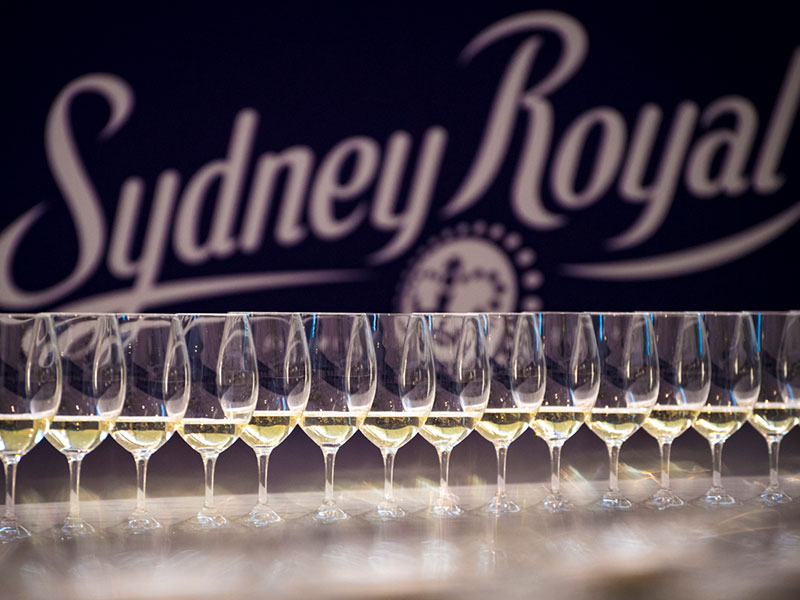 Wine glasses filled with white wine samples lined up in front of a blurred Sydney Royal sign at the Sydney Royal Wine Show.
