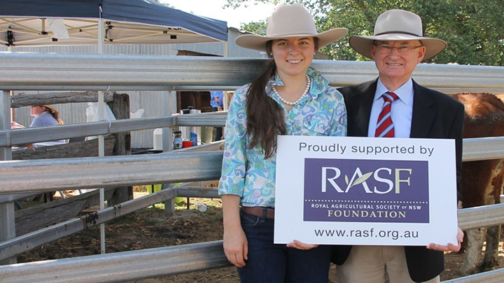 Two people in hats standing by livestock yards, holding a sign supporting the RAS Foundation.