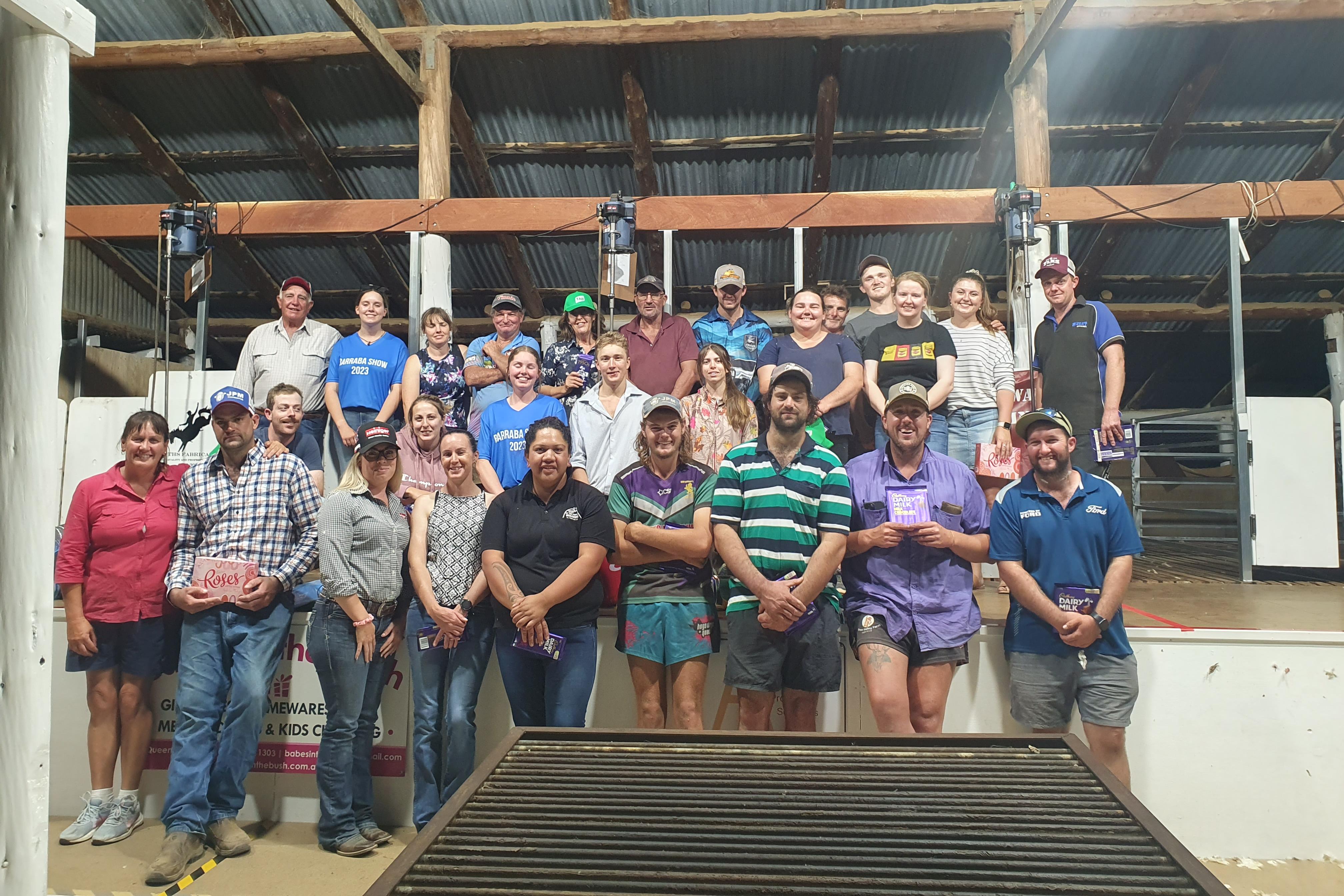 Large group of people standing together inside a rural shed, smiling and posing for a group photo.
