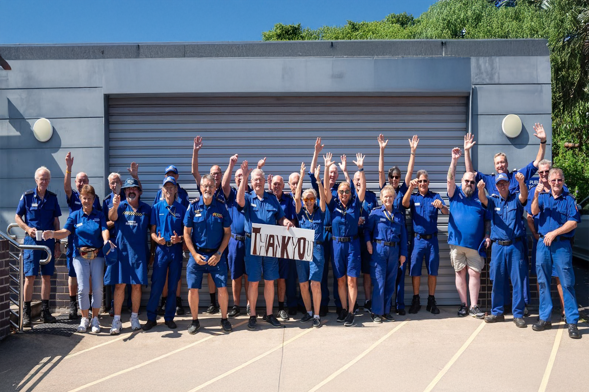 Group of uniformed marine rescue volunteers in Ulladulla, smiling and waving while holding a “Thank You” sign outside a building.
