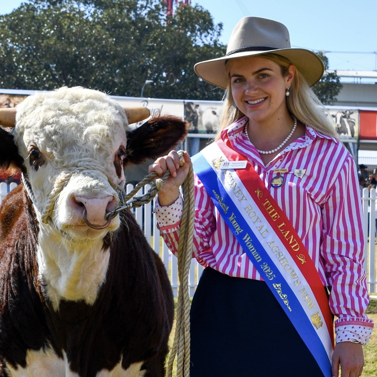 Cassandra Bush, a woman with blonde hair, smiling in a striped shirt and hat holding a Hereford bull, wearing Sydney Royal AgShows NSW winner sashes.