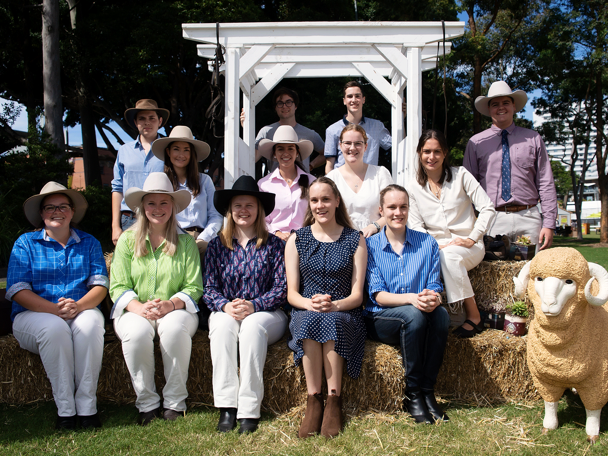 The RAS Rural Scholaship recipients, smiling and wearing rural attire and hats, seated on hay bales outdoors with a ram statue.