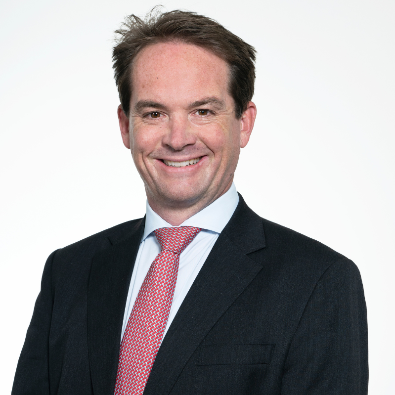 Headshot of Treasurer Dr. Sam Walker, smiling and  wearing a dark suit, white shirt, and red patterned tie.