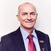 Headshot of Chair of Woodchop Stephen Dadd, smiling and wearing a dark suit jacket, light blue checked shirt, and red tie.