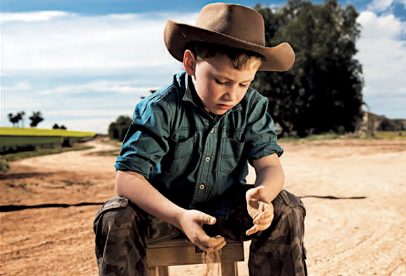A boy in a hat and green shirt sitting on a wooden stool examining soil on a dirt track.