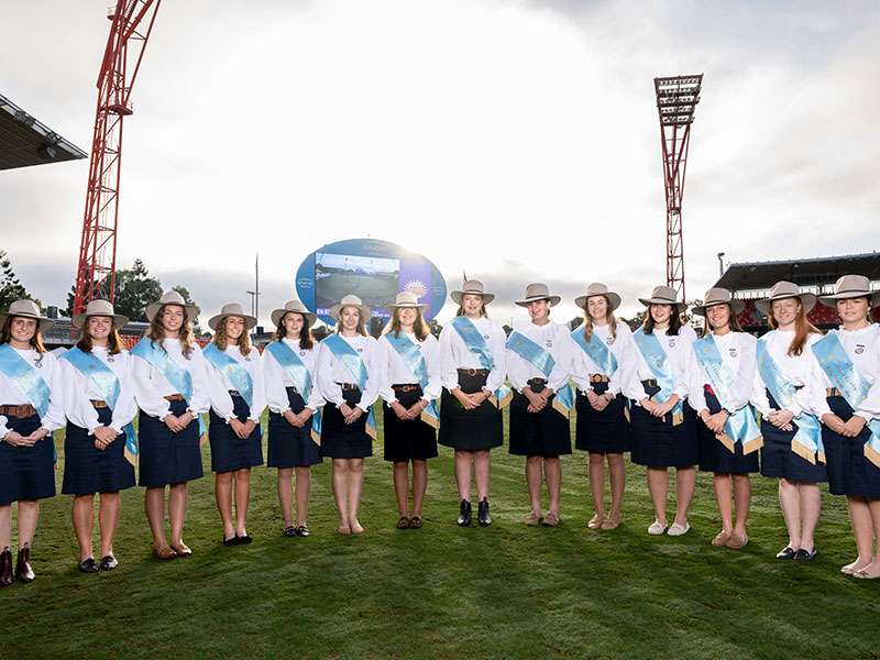 A group of young women standing and lined up in white blouses, navy skirts, light blue sashes and Akubra hats, smiling in the middle of ENGIE Stadium.
