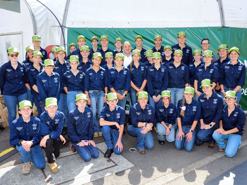 High school students wearing navy shirts and fluoro green hats, smiling together with their teachers at an All Roads to the Royal excursion.