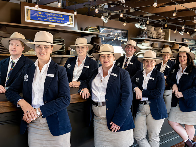 Eight finalists of the 2026 RAS Rural Achiever Award wearing white shirts, navy blazers and tan Akubra hats, leaning on a counter at a hat shop and smiling.