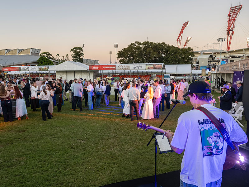 Crowds of people are socialising on the Cattle Judging Lawn while a man performs on stage at the Hats Off to the Royal Event.