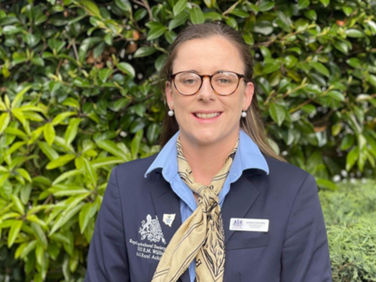 RAS Youth Group member Jessica Fearnley wearing glasses and a navy blazer with an RAS badge, smiling against green foliage.