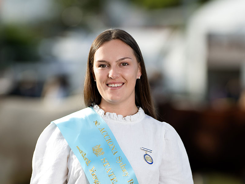 Headshot of 2026 Young Woman finalist Emily Greer, a woman with long dark hair, wearing a white shirt and a light blue sash.