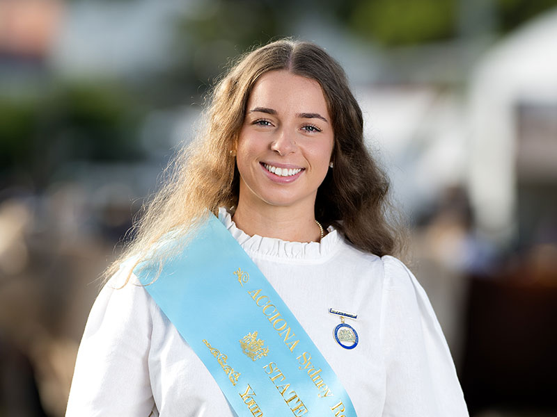 Headshot of 2026 Young Woman finalist Libby Coles, a brunette woman with long wavy hair, wearing a white shirt and a light blue sash.
