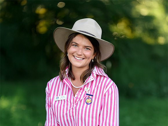 Headshot of 2025 finalist Tessa Parry, a woman with brown wavy hair, smiling with pearl drop earrings and a wide-brimmed hat.