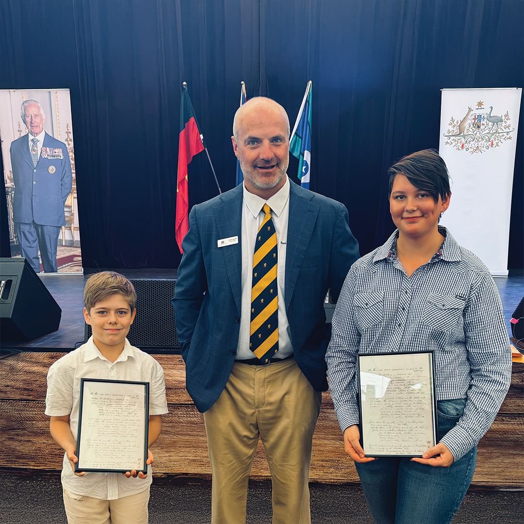Two students holding framed certificates standing with Duncan Kendall, on stage with Australian flags behind them.