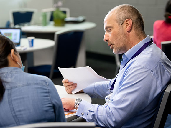 A man in a blue shirt holding papers while discussing with a colleague in a classroom for Teachers Professional Development Day.