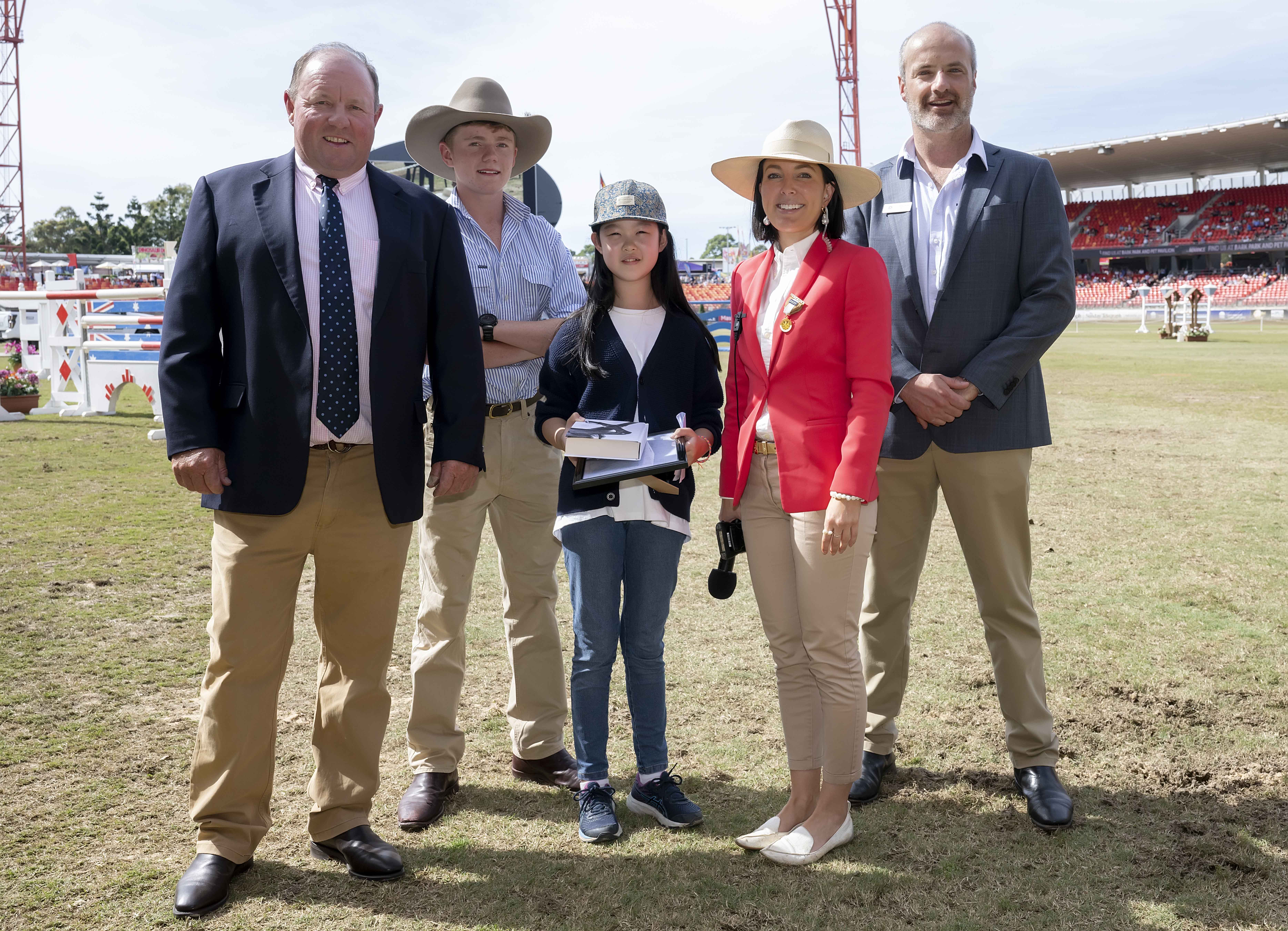 A young student holding certificates with officials and presenters smiling beside her on a showground field, presented with the 2022 RAS AB Paterson Bush Poetry Award.