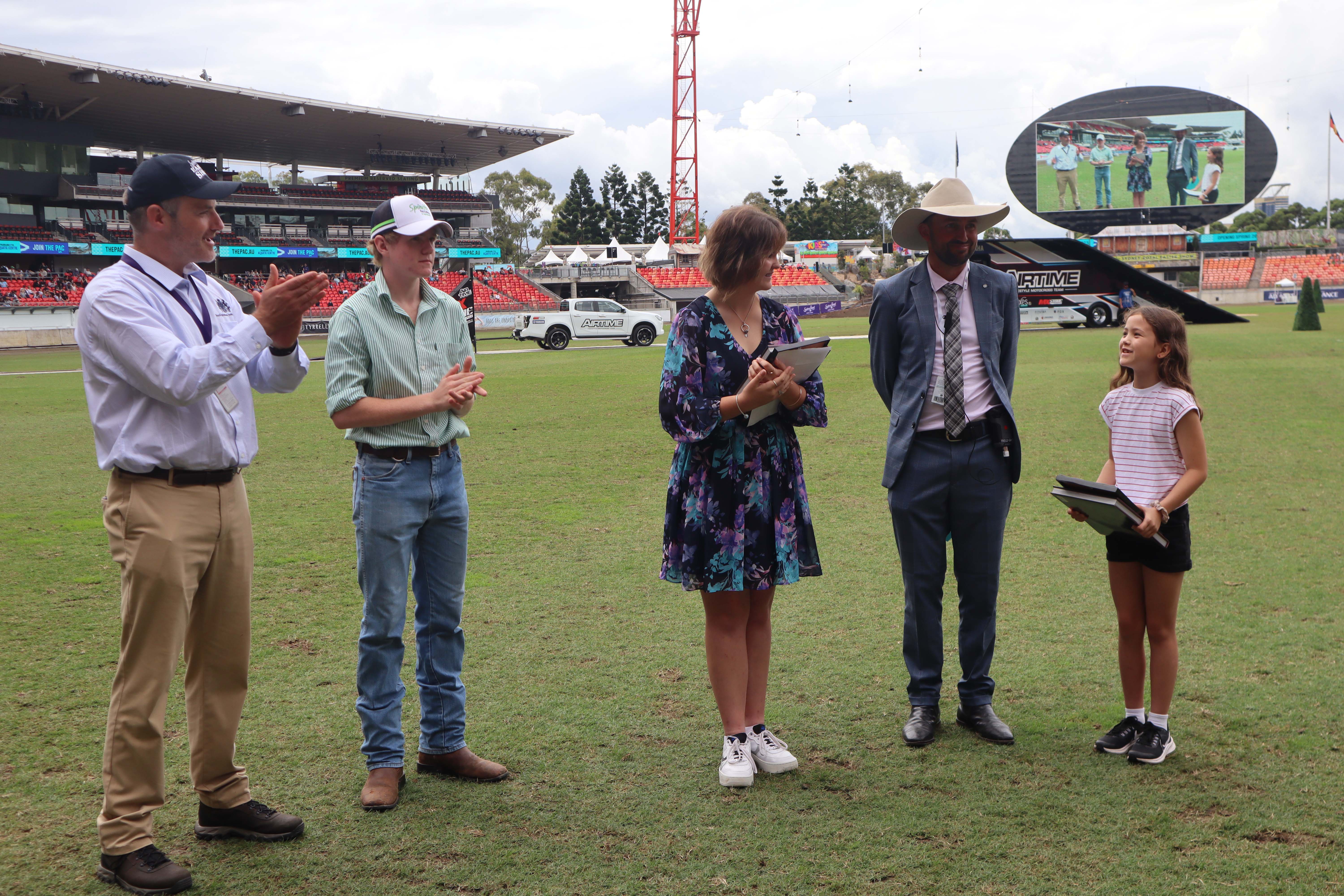 Four adults and a student on a stadium field applauding the Primary School winner of the 2023 RAS AB Paterson Bush Poetry Award.