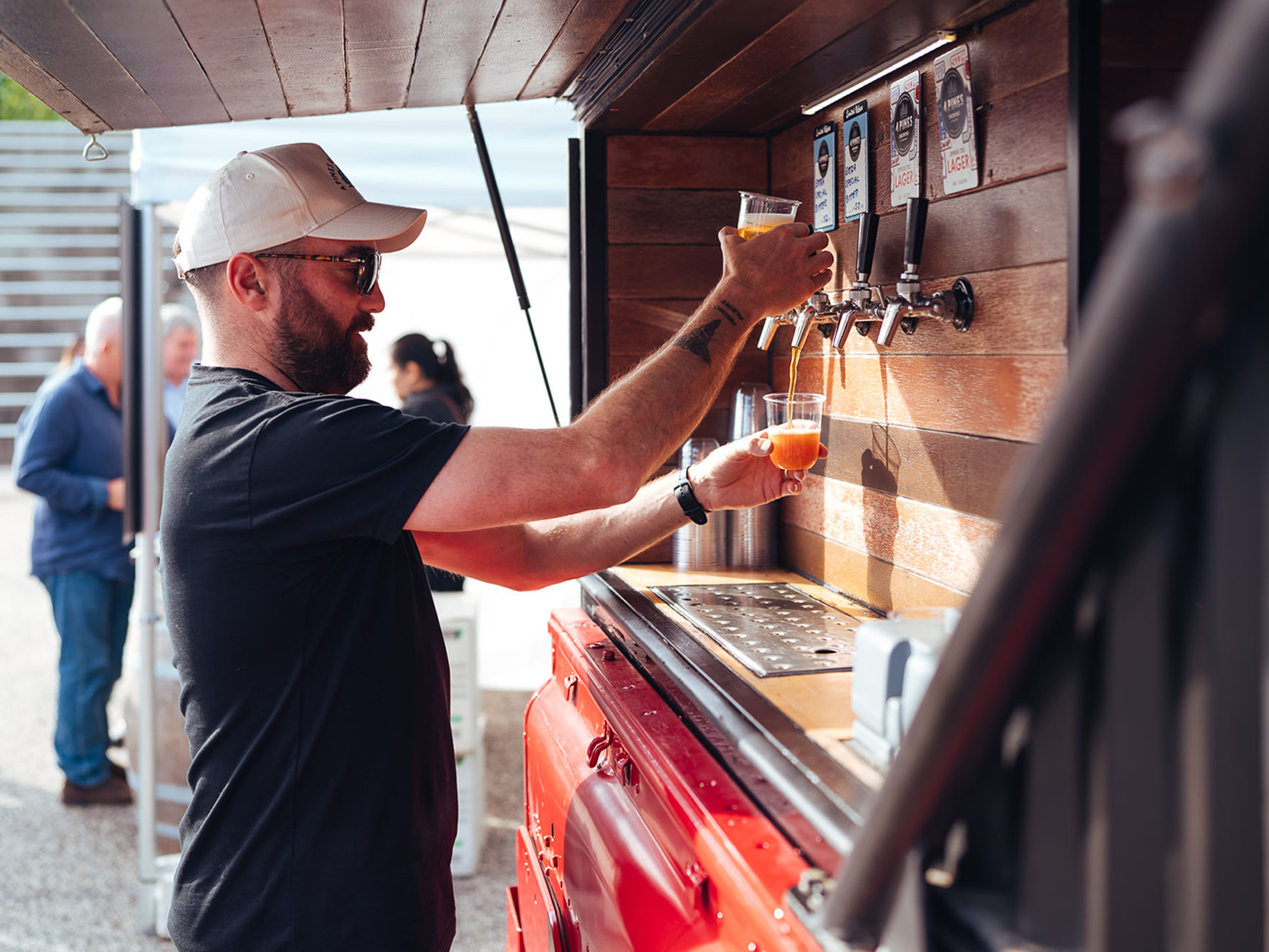 A patron wearing a cap pours a glass of 4 Pines Beer from a tap at an outdoor bar.