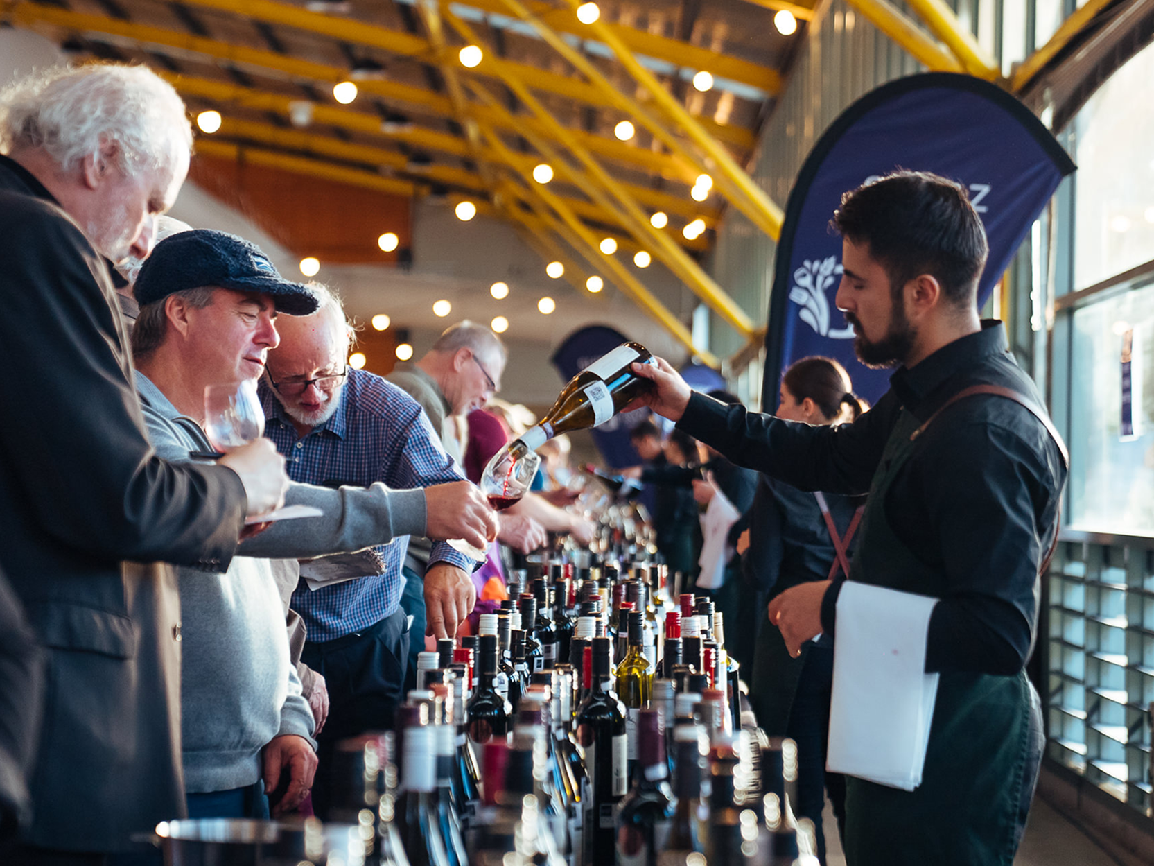 Guests at a Grape, Grain and Graze Festival event sample wine as a server pours their glasses, inside a hall with yellow beams.