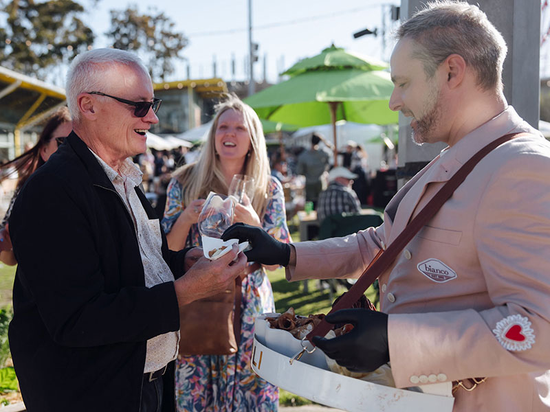 A waiter in a dusty pink shirt serving cannoli to a smiling man in a black jacket and glasses at the Grape Grain and Graze Festival.