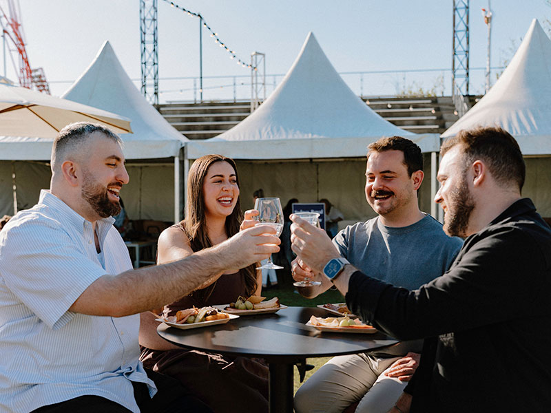 A group of people smiling and toasting their glasses at a table at the Grape Grain and Graze Festival.