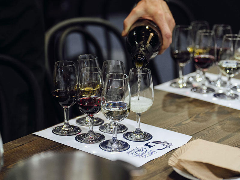 A bottle of clear distilled spirits poured into a sampling glass with five different glasses filled with alcohol at the Grape Grain and Graze Festival.
