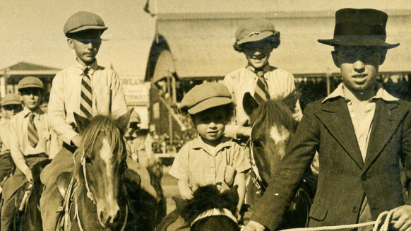 Sepia photograph of children wearing hats and ties riding ponies at a showground.