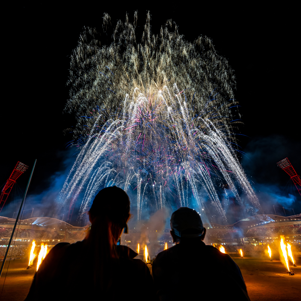 Two people silhouetted against a night sky watching a vibrant fireworks display in a stadium.