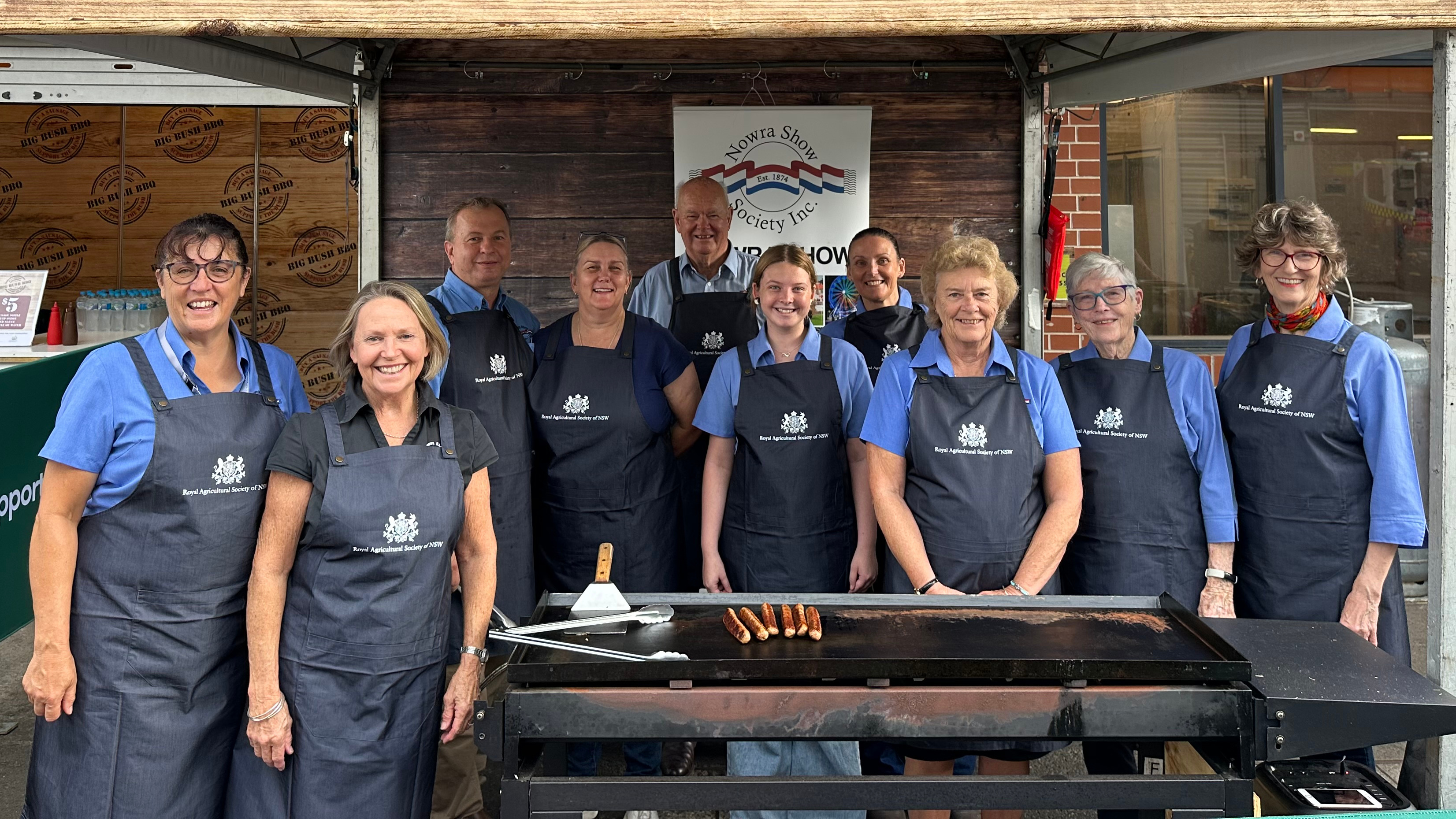 Group of Big Bush BBQ  volunteers wearing navy aprons with Royal Agricultural Society logos stand smiling behind a barbecue at a show stall.