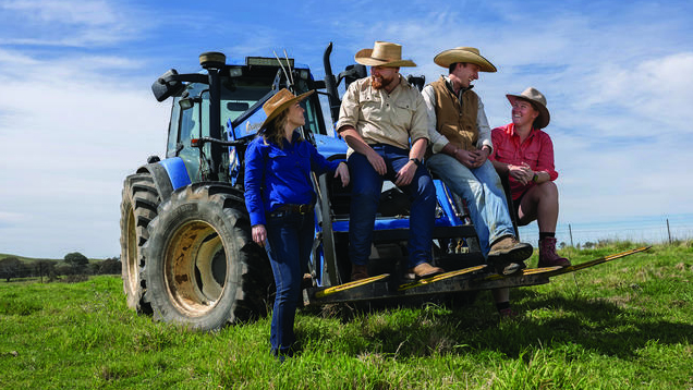 Four people wearing hats sit together on a blue tractor in a grassy paddock under blue sky.